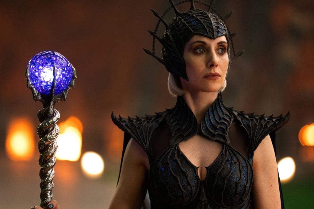 A woman in elaborate black armor and a spiked helmet holds a glowing blue staff, standing against a blurred background with warm lights.