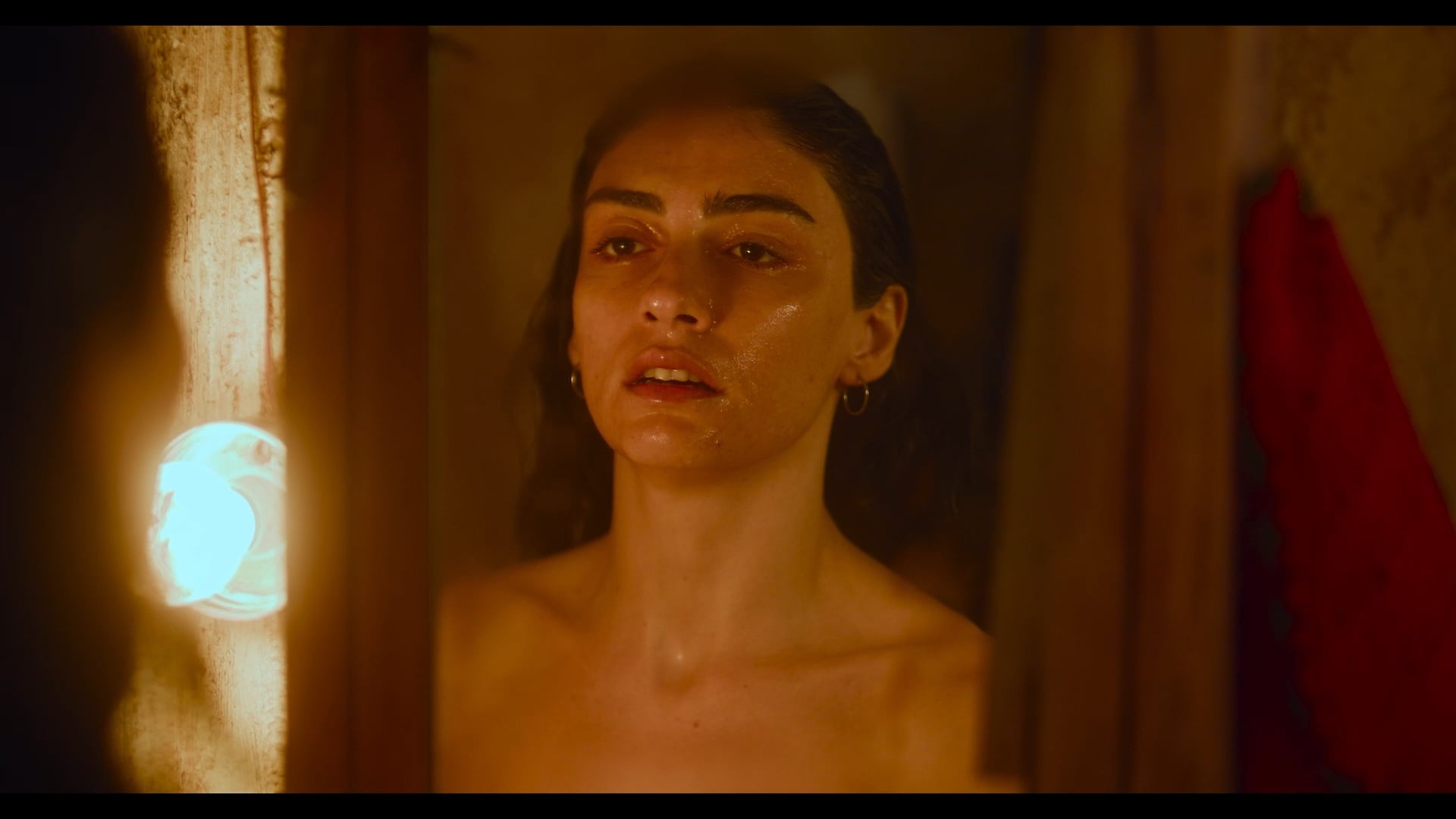 A woman with wet hair and dewy skin looks at her reflection in a mirror, illuminated by warm light from a bulb on the left side.