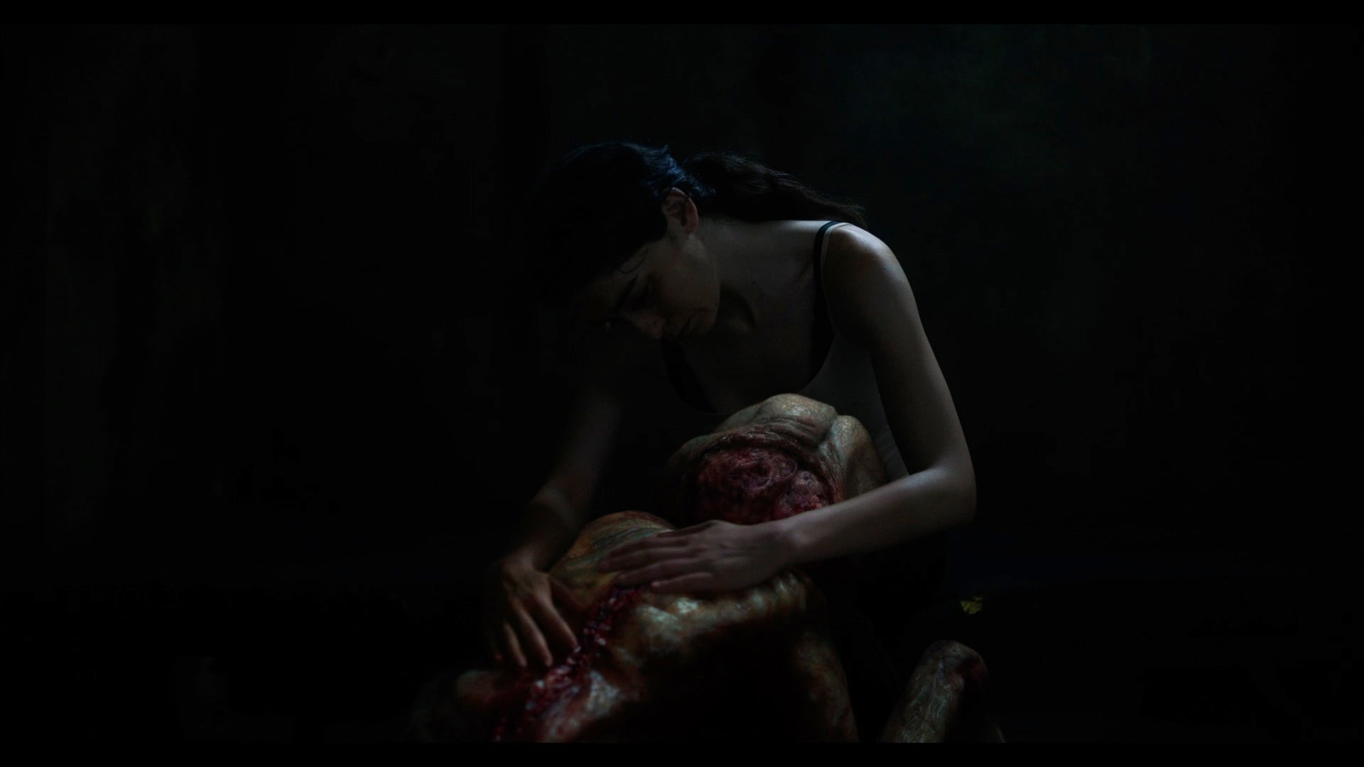 A woman in a dark room tends to a large, grotesque, partially skinned creature with exposed flesh and muscle.