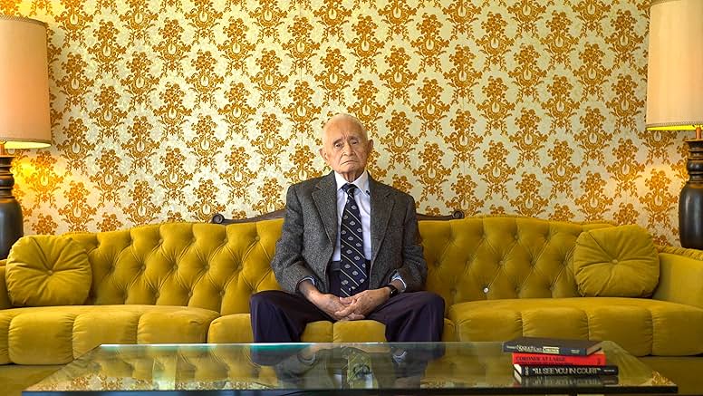 An older man in a suit sits on a yellow tufted sofa in front of patterned wallpaper. A glass coffee table with books is in front of him, and lamps are on either side of the sofa.