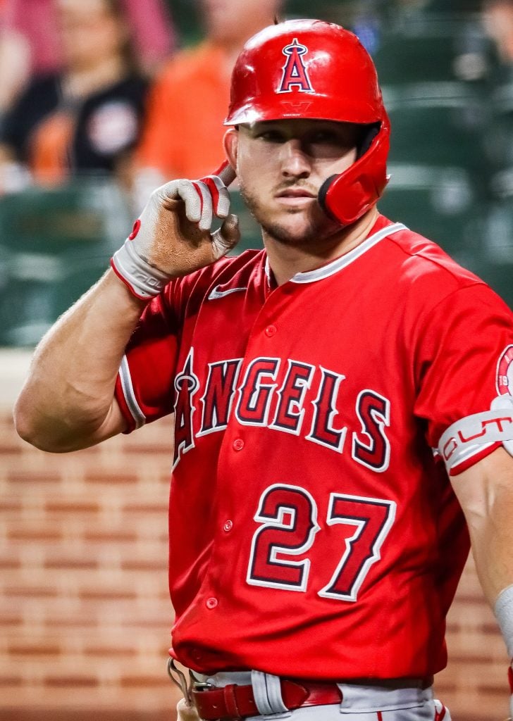 A baseball player in an Angels uniform with the number 27 stands on the field, wearing a red helmet and holding his batting glove to his face.