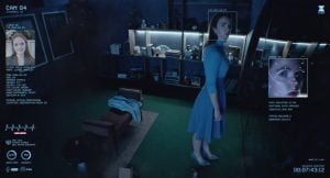 A woman in a blue dress stands in a dimly lit room, viewed by a surveillance camera interface displaying identification and facial recognition data.