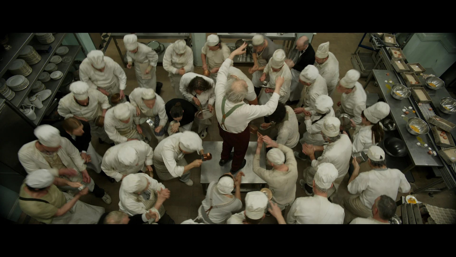 A group of chefs in white uniforms surrounds a man standing on a table, seen from overhead in a busy kitchen setting.