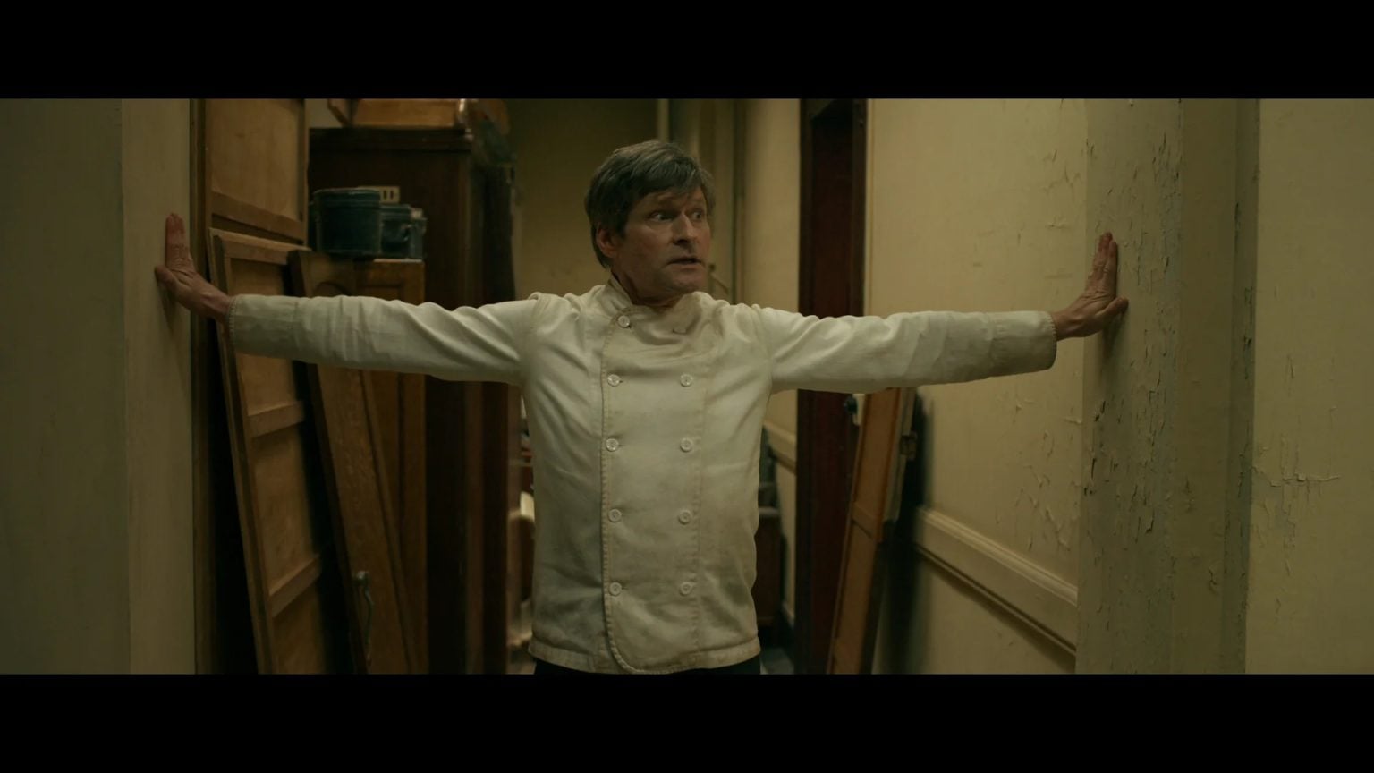 A chef in a white jacket stands in a narrow hallway with arms outstretched, touching both walls and looking to the side.