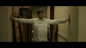 ‘Mr. K’ Blu-Ray Review – Trippy Psychological Spiral Is An Intriguing Relinquishing Of Control A chef in a white jacket stands in a narrow hallway with arms outstretched, touching both walls and looking to the side.
