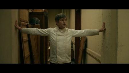 A chef in a white jacket stands in a narrow hallway with arms outstretched, touching both walls and looking to the side.