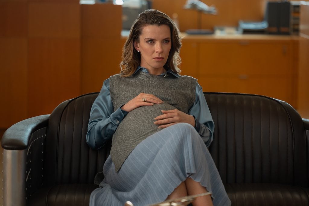 A woman in a blue blouse and gray vest sits on a dark couch, resting her hands on her pregnant belly, looking forward with a serious expression.