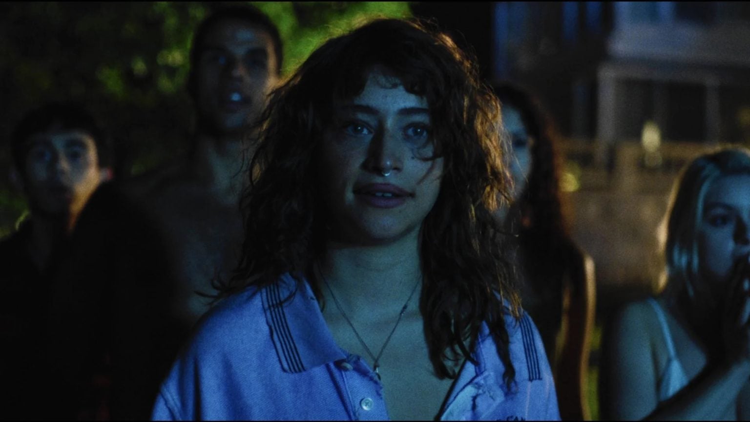 A woman with curly hair and a nose ring stands in front of a group of people outdoors at night, looking ahead with a neutral expression.