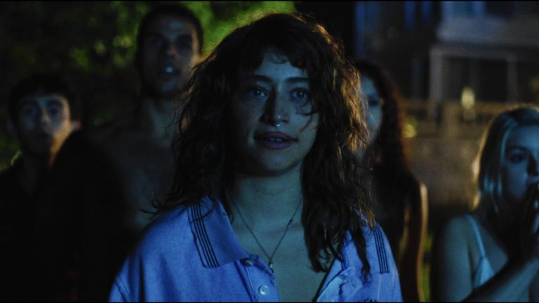 A woman with curly hair and a nose ring stands in front of a group of people outdoors at night, looking ahead with a neutral expression.