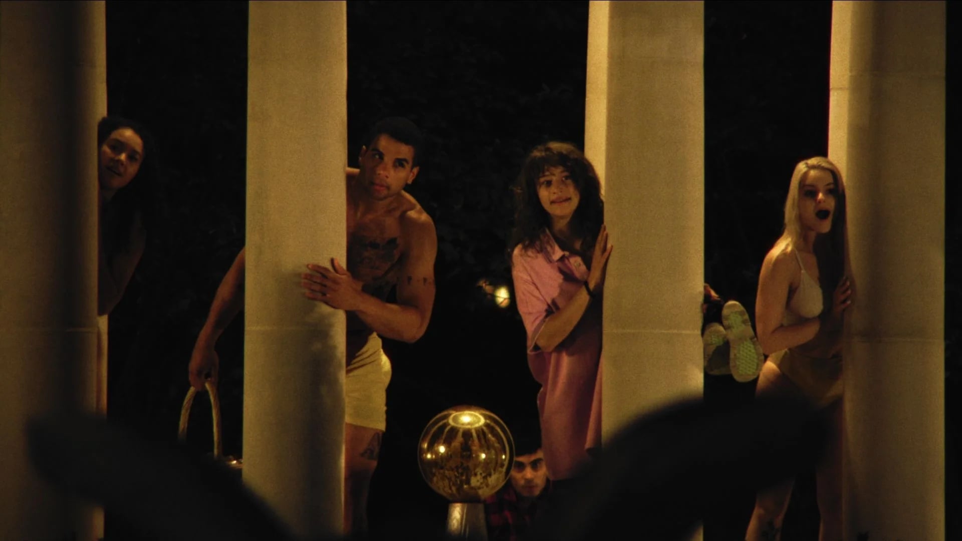 Four people stand behind large columns at night, peeking out and looking forward; two are in swimwear, one in a robe, and one in a towel, with a golden decorative object in front of them.