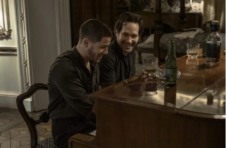 ‘Power Ballad’ Review – This Paul Rudd & Nick Jonas Comedy Strikes An Emotional Chord Two men sit at a piano, smiling and laughing. There are drinks and a pair of sunglasses on top of the piano in a warmly lit room.