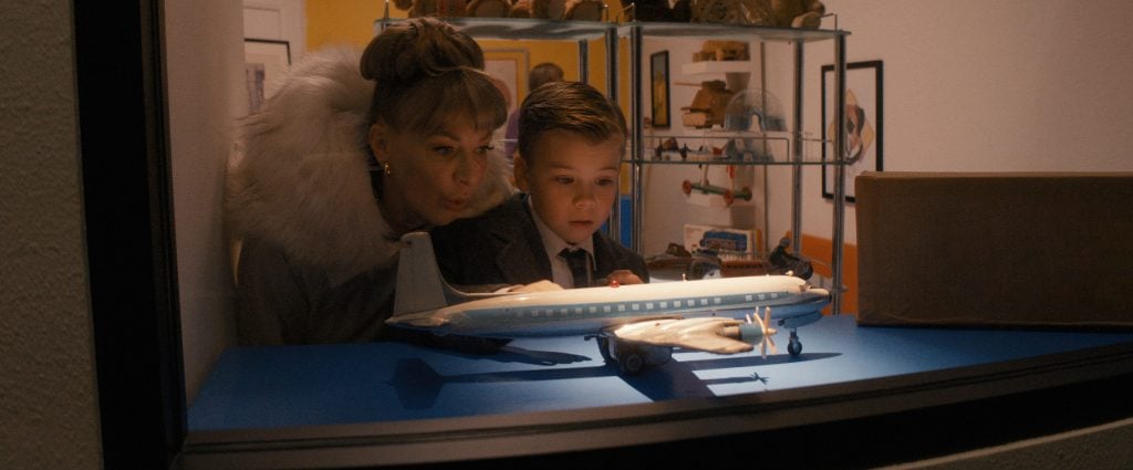 ‘Propeller One-Way Night Coach’ Review – A Memory As A Movie A woman and a young boy stand behind glass, closely observing a model airplane displayed on a blue surface, with shelves of collectibles in the background.