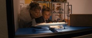 ‘Propeller One-Way Night Coach’ Review – A Memory As A Movie A woman and a young boy stand behind glass, closely observing a model airplane displayed on a blue surface, with shelves of collectibles in the background.