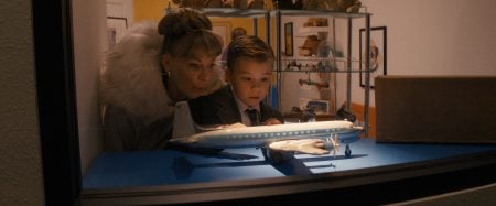 ‘Propeller One-Way Night Coach’ Review – A Memory As A Movie A woman and a young boy stand behind glass, closely observing a model airplane displayed on a blue surface, with shelves of collectibles in the background.