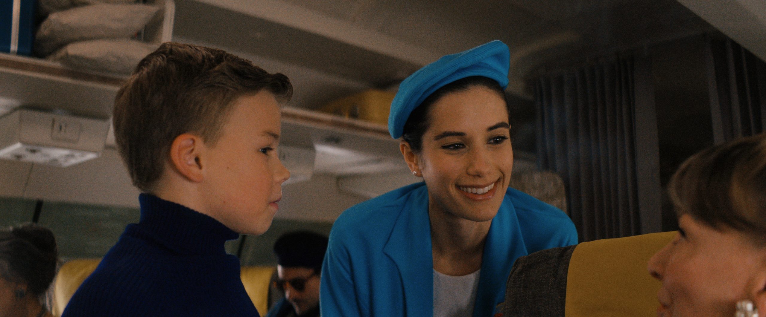 A flight attendant in a blue uniform smiles while talking to a seated woman and a young boy in a blue sweater on an airplane.