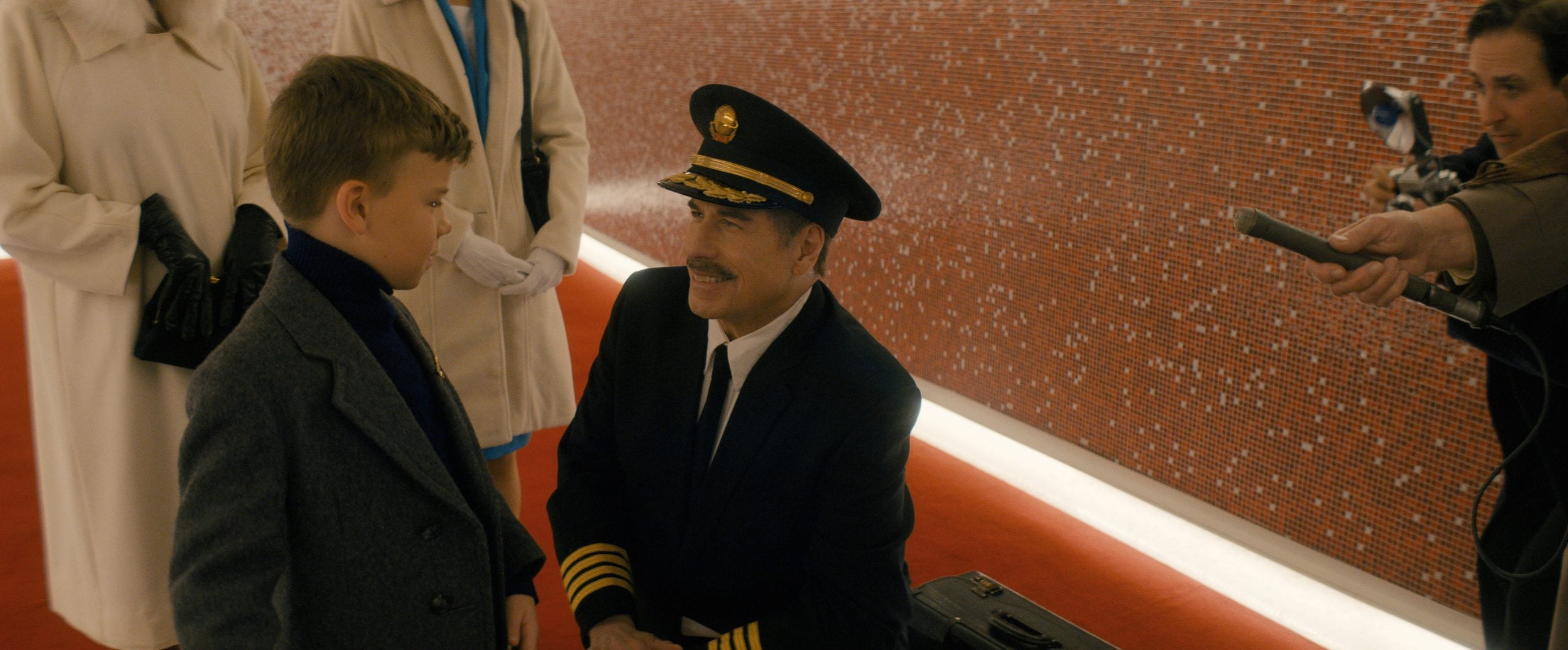 A man in a pilot's uniform kneels to speak to a young boy in a coat, while adults stand nearby and a cameraman films the scene in a red-carpeted hallway.