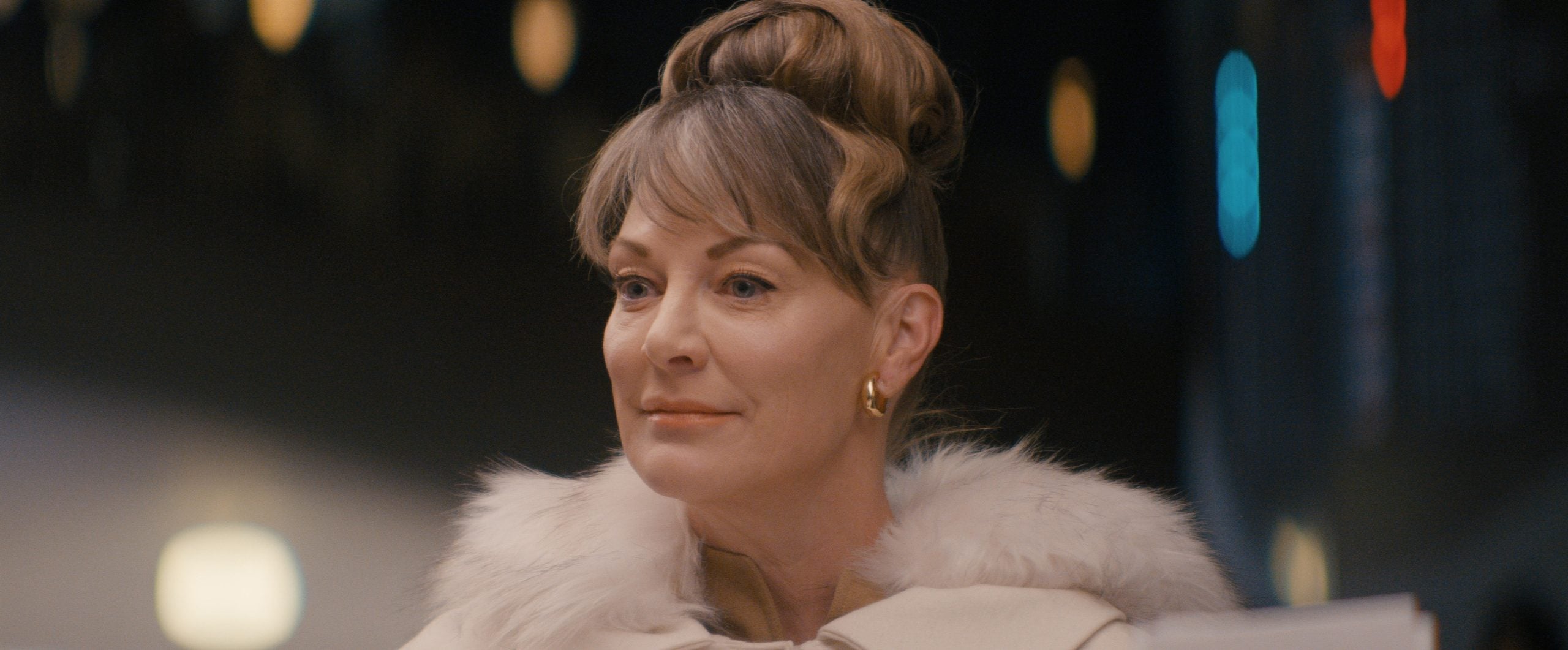 A woman with light brown hair styled in an updo, wearing gold hoop earrings and a white fur-collared coat, looks ahead with a neutral expression.