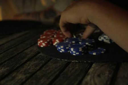 A hand reaches for a stack of poker chips, with red and blue chips arranged on a round black mat atop a wooden table.