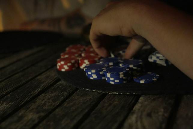 A hand reaches for a stack of poker chips, with red and blue chips arranged on a round black mat atop a wooden table.