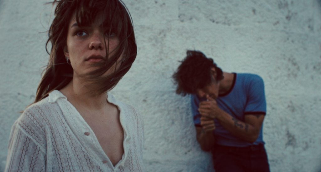 A woman with windblown hair stands in the foreground, looking away, while a man in a blue shirt leans against a white wall in the background, lighting a cigarette.