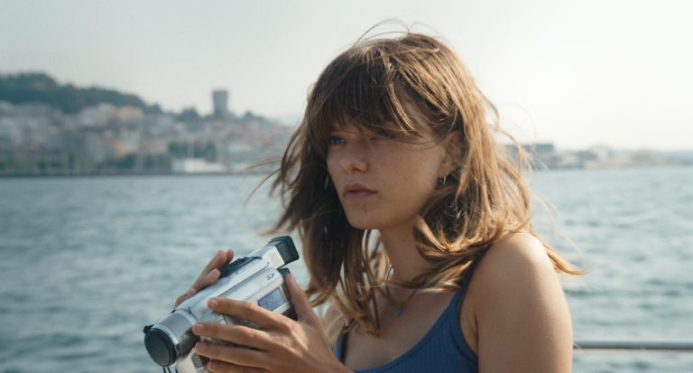 A woman with tousled hair holds a handheld video camera while standing near water, with a cityscape visible in the background.