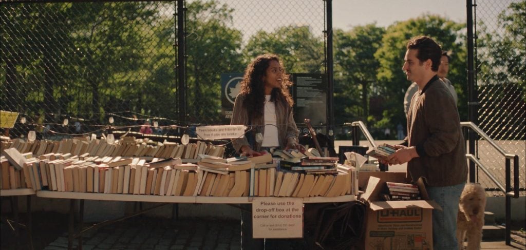 Two people stand at an outdoor table filled with stacks of books for sale or donation, with a fence and trees in the background.