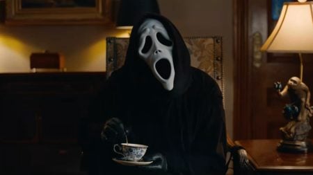 A person wearing a white Ghostface mask and black hooded robe sits in an armchair, holding and stirring a teacup.