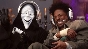 Two people wearing headsets play video games on a couch; one wears a Ghostface mask and black robe, while the other laughs, holding a controller with a bandaged hand.