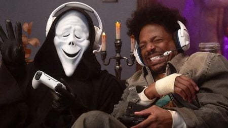 Two people wearing headsets play video games on a couch; one wears a Ghostface mask and black robe, while the other laughs, holding a controller with a bandaged hand.