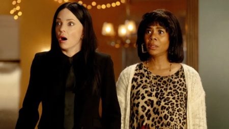 Two women stand side by side indoors, both looking ahead with surprised expressions. One wears black; the other wears a leopard print top and a white cardigan.