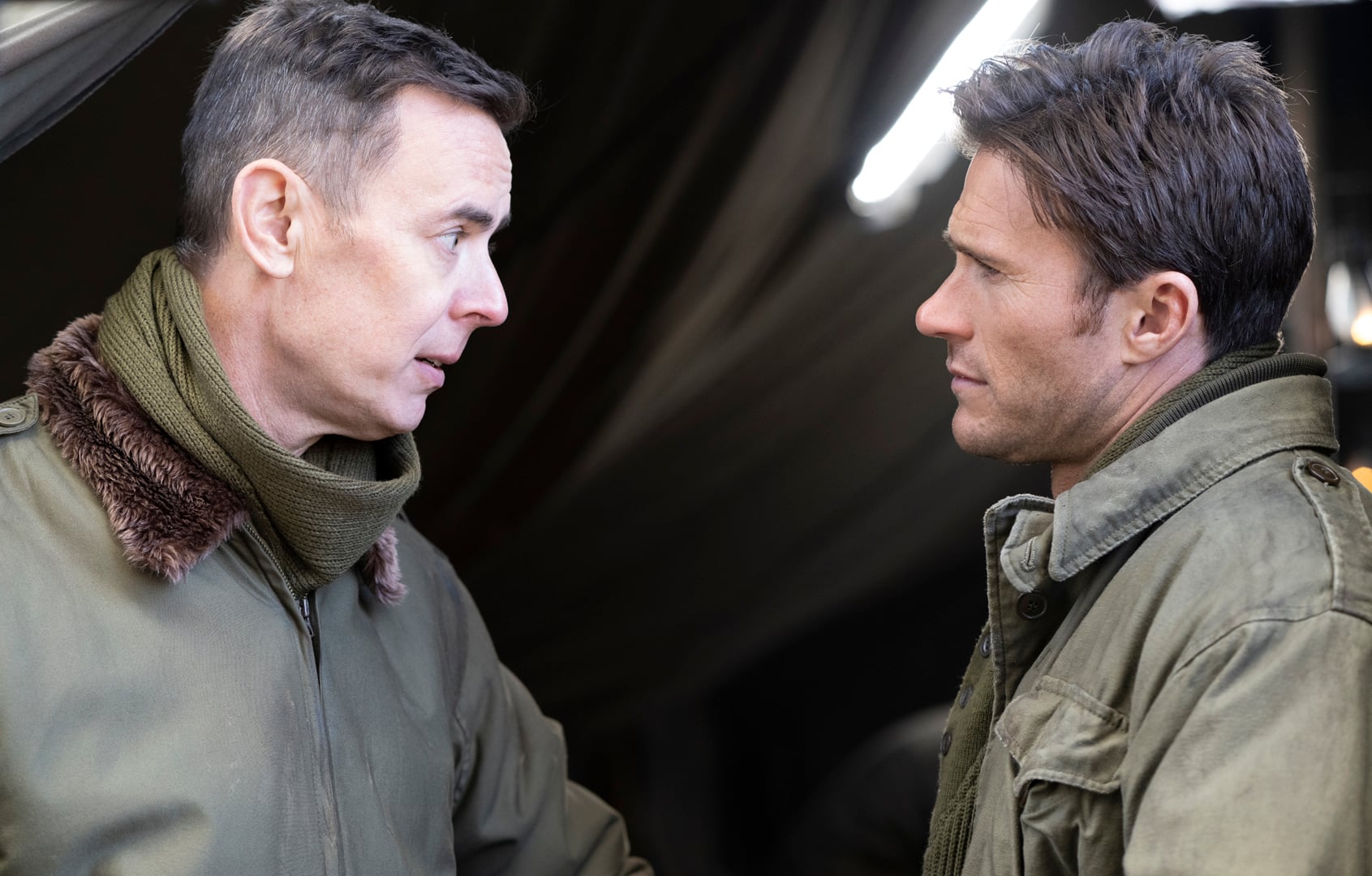 Two men in military-style jackets stand facing each other indoors, engaged in a serious conversation.