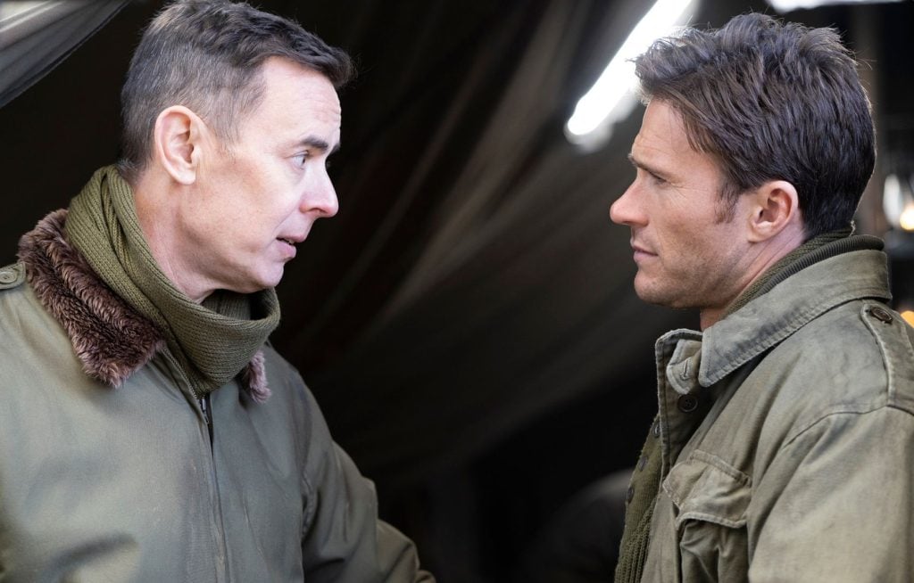 Two men wearing military-style jackets stand facing each other indoors, appearing to have a serious conversation.