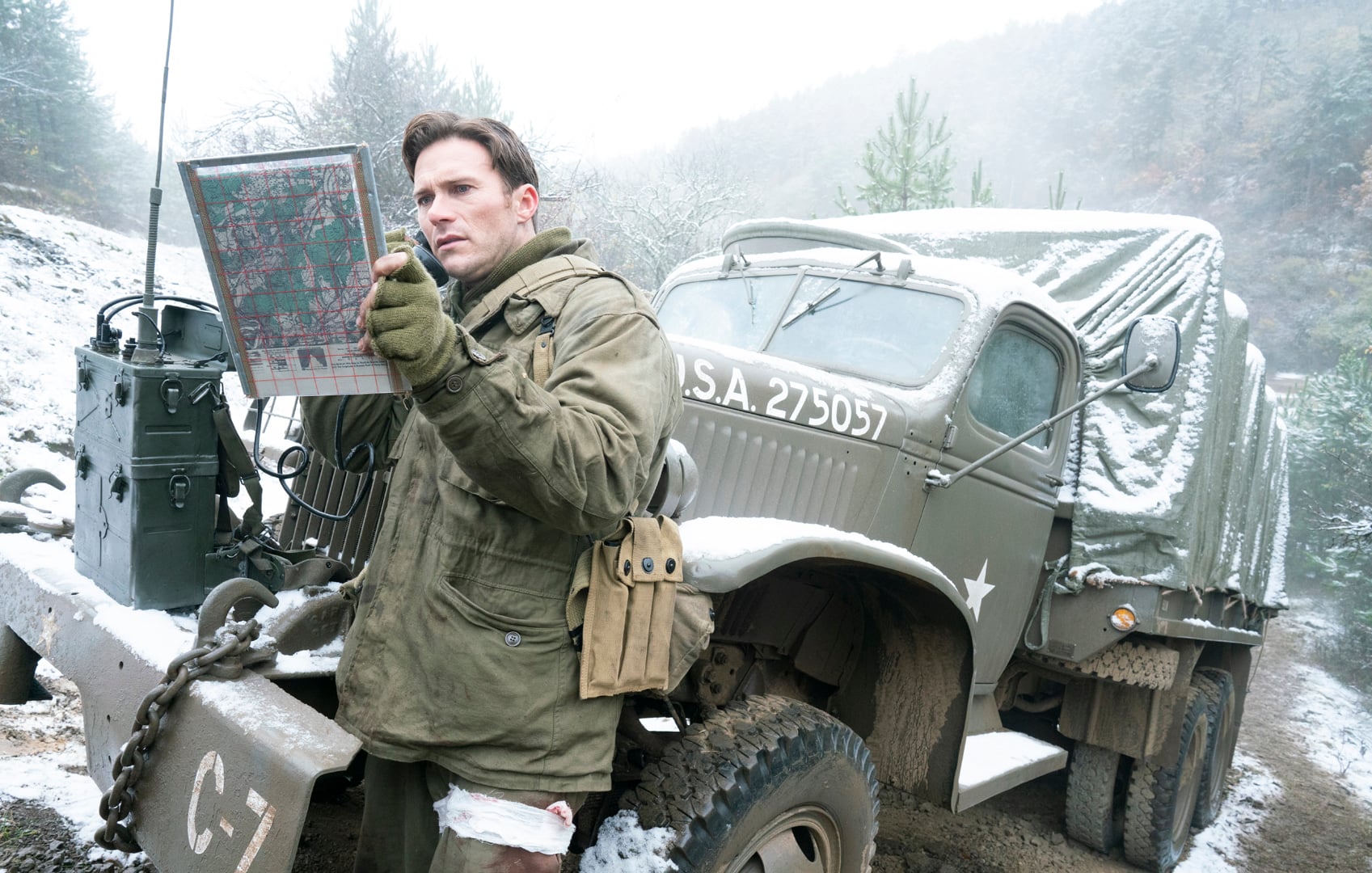 A soldier in winter gear examines a map in front of a military truck marked "U.S.A. 275057" on a snowy roadside with trees and hills in the background.