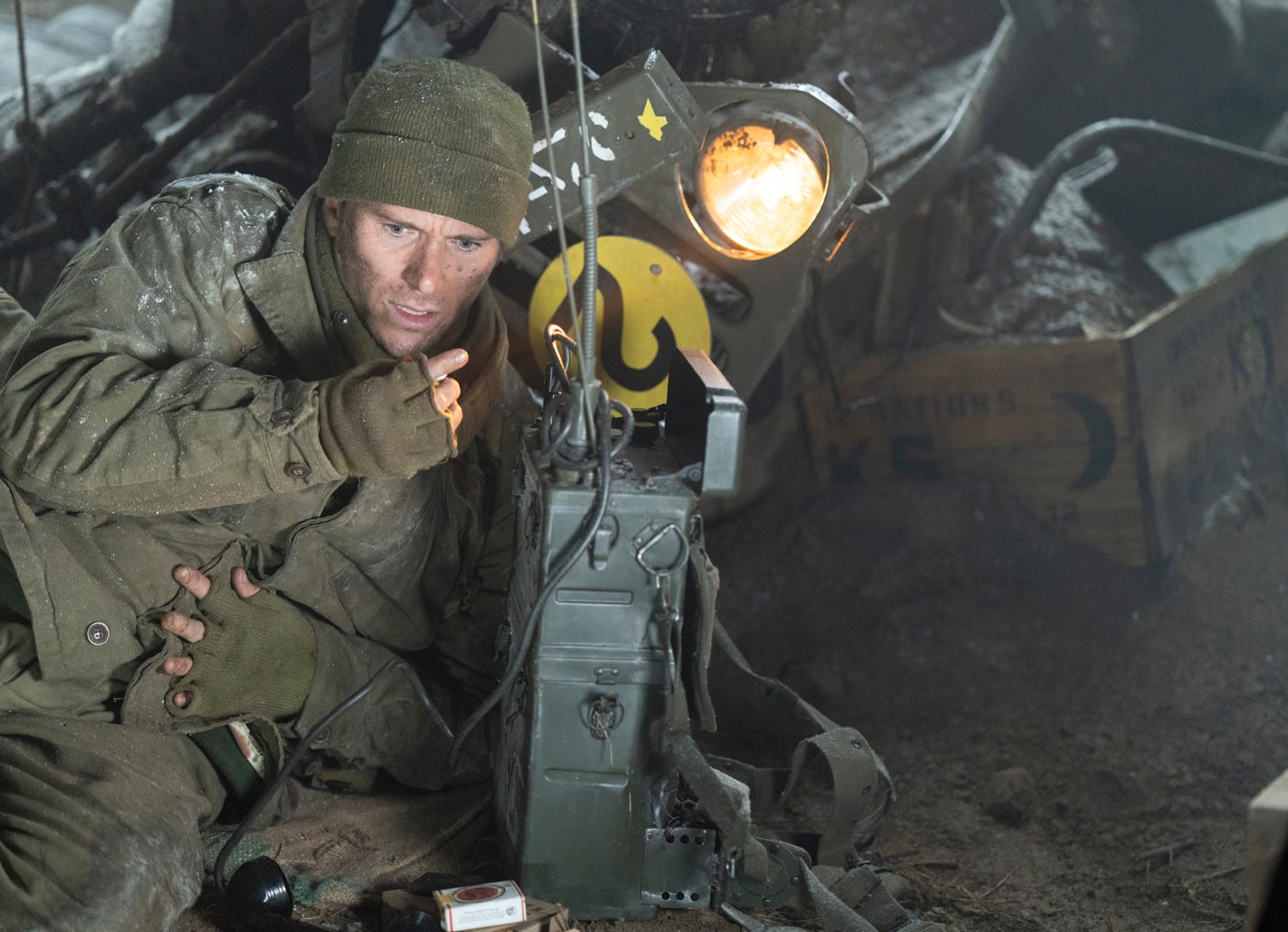 A soldier in winter gear crouches next to a military radio, holding a lit cigarette, with a tank and wooden crate in the background.