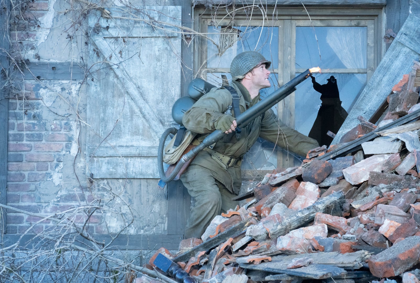 A soldier in World War II-era gear climbs over rubble, aiming a rifle out of a broken window in a damaged brick building.