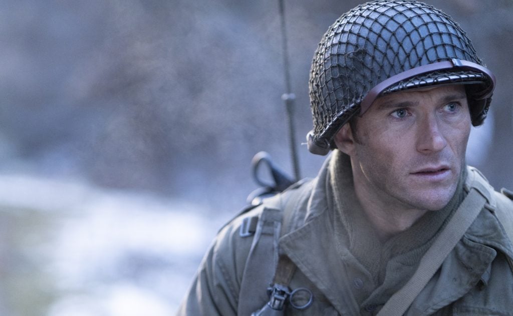 A soldier wearing a helmet and military uniform looks to the right; the background is blurred and outdoors.