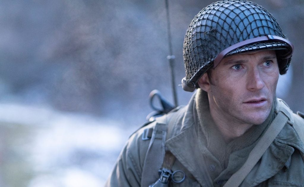 A man dressed as a World War II soldier wears a helmet and military gear, looking intently to the side outdoors in a cold, misty setting.