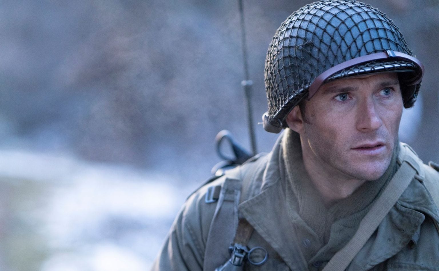 A man dressed as a World War II soldier wears a helmet and military gear, looking intently to the side outdoors in a cold, misty setting.
