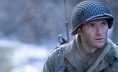 A man dressed as a World War II soldier wears a helmet and military gear, looking intently to the side outdoors in a cold, misty setting.