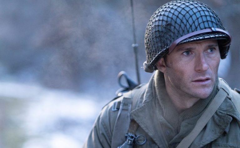 A man dressed as a World War II soldier wears a helmet and military gear, looking intently to the side outdoors in a cold, misty setting.
