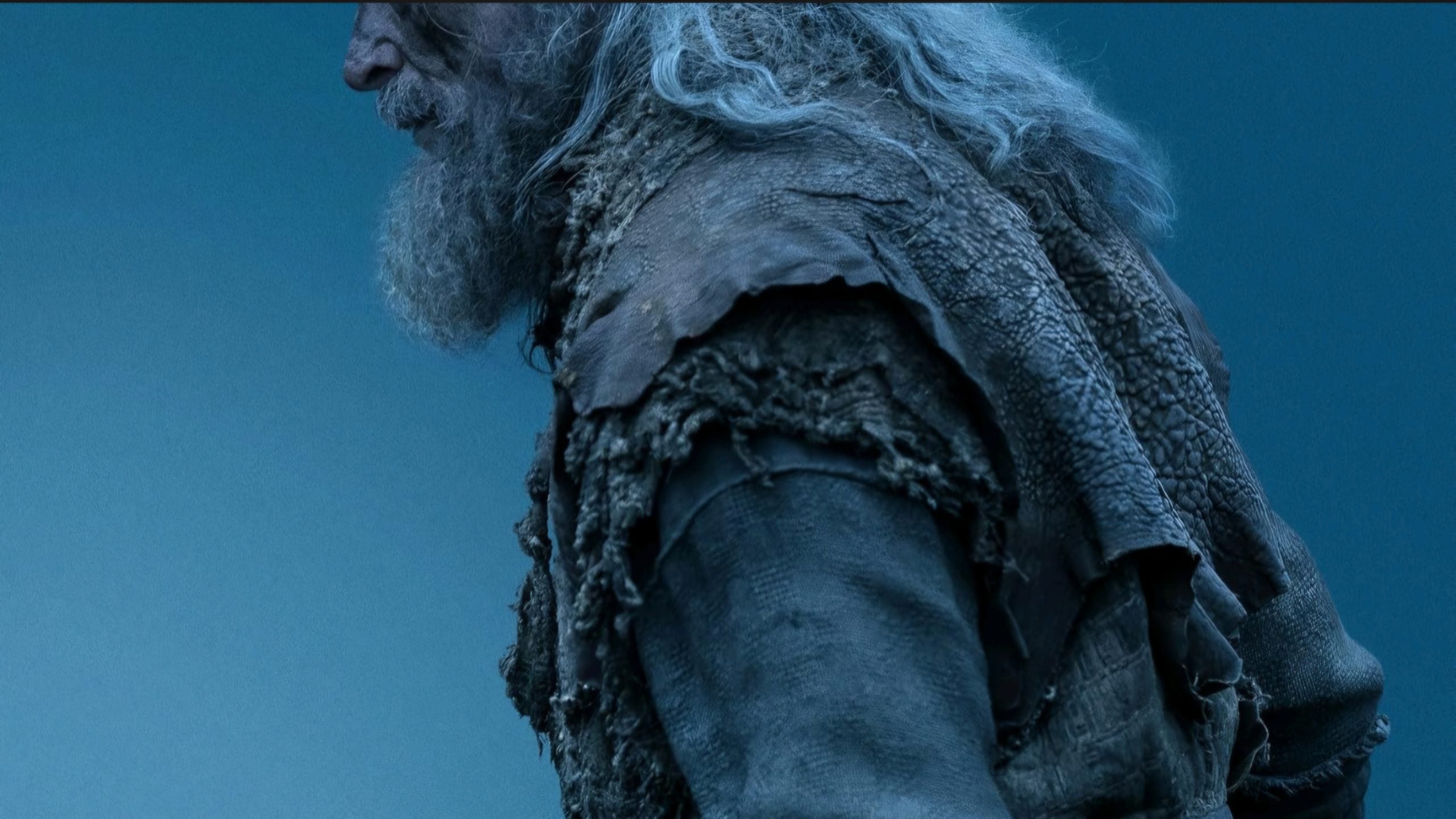 A person with long gray hair and a beard wears layered, weathered clothing made of rough fabric and leather, shown against a blue background.