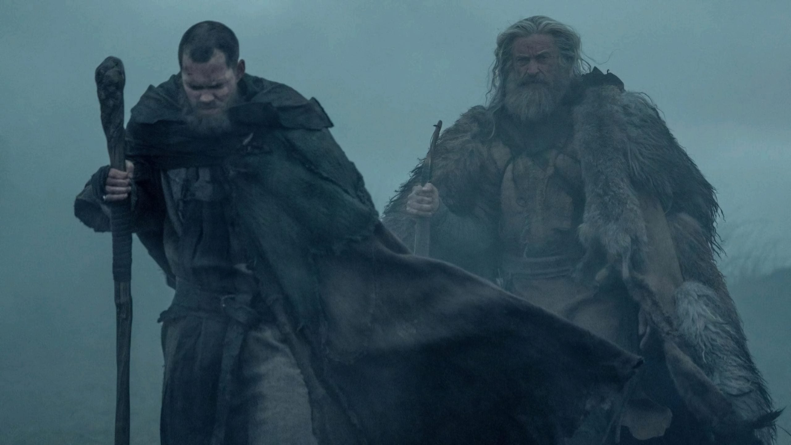 Two men in heavy cloaks walk through dense fog, one holding a staff and the other wearing fur, in a cold, rugged outdoor setting.