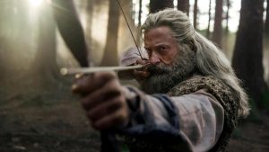 ‘The Death of Robin Hood’ Review: Violent, Dull, And Stagnant A man with long gray hair and a beard draws a bow and aims an arrow in a dense forest, wearing rustic clothing.