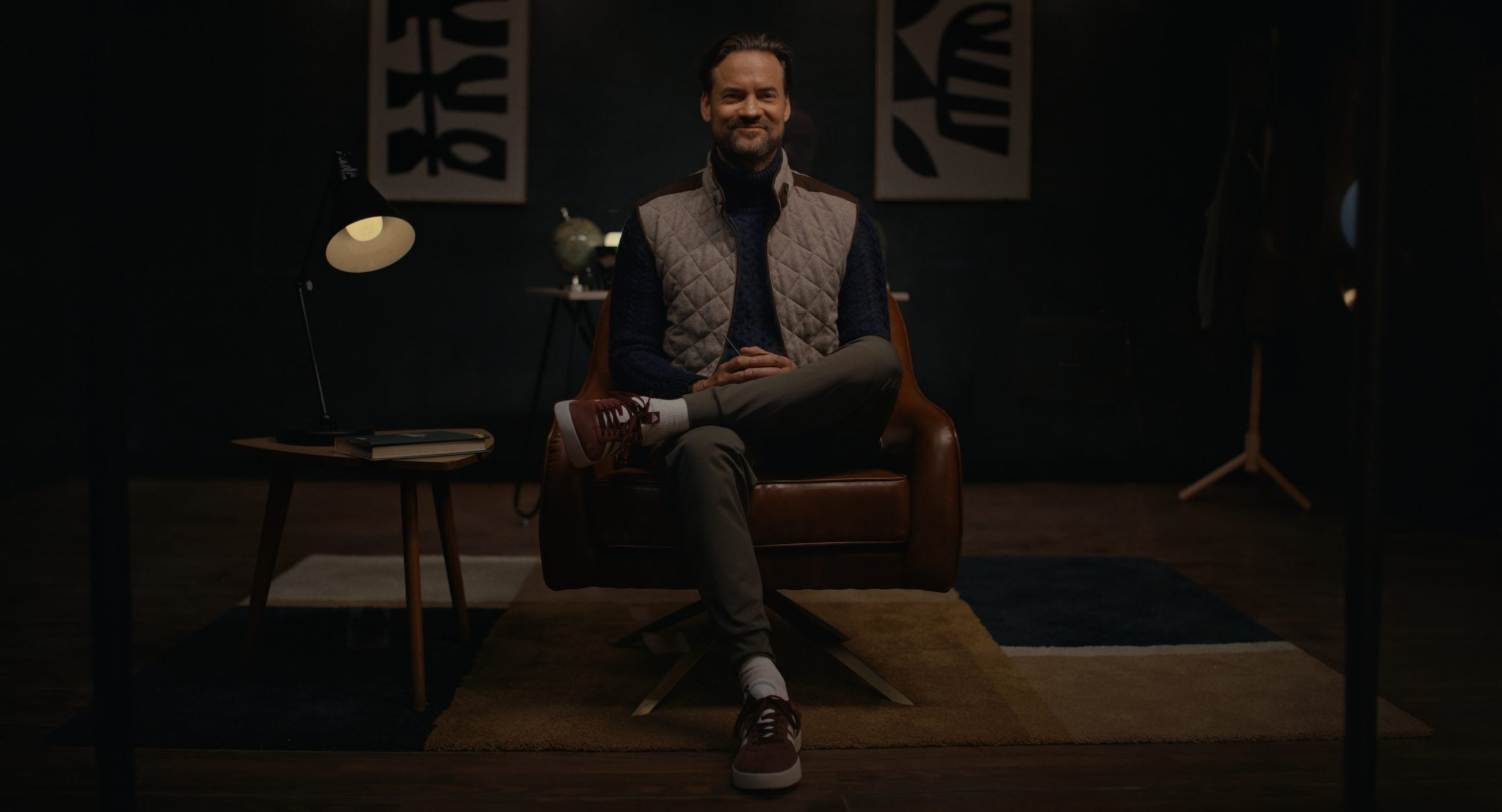 A man sits cross-legged in a brown leather chair, smiling at the camera. A small table with a lamp and books is beside him, with abstract art on the dark wall behind.