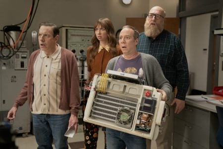 Four people stand in a lab, looking surprised; one person in front holds a large machine with exposed wires and components.