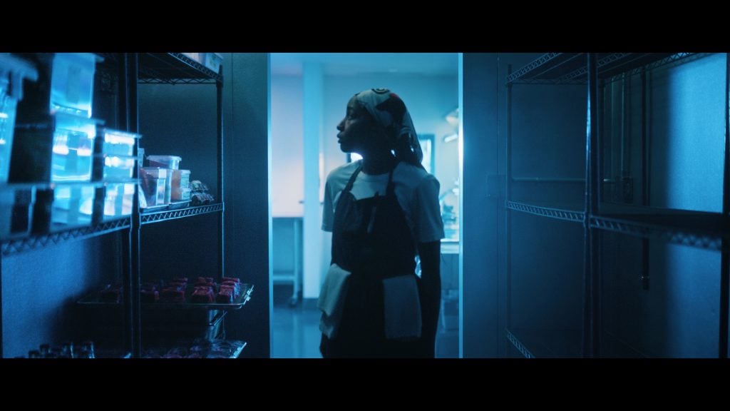 A person wearing an apron and headscarf stands in a dimly lit storage room with metal shelves, illuminated by blue lighting.