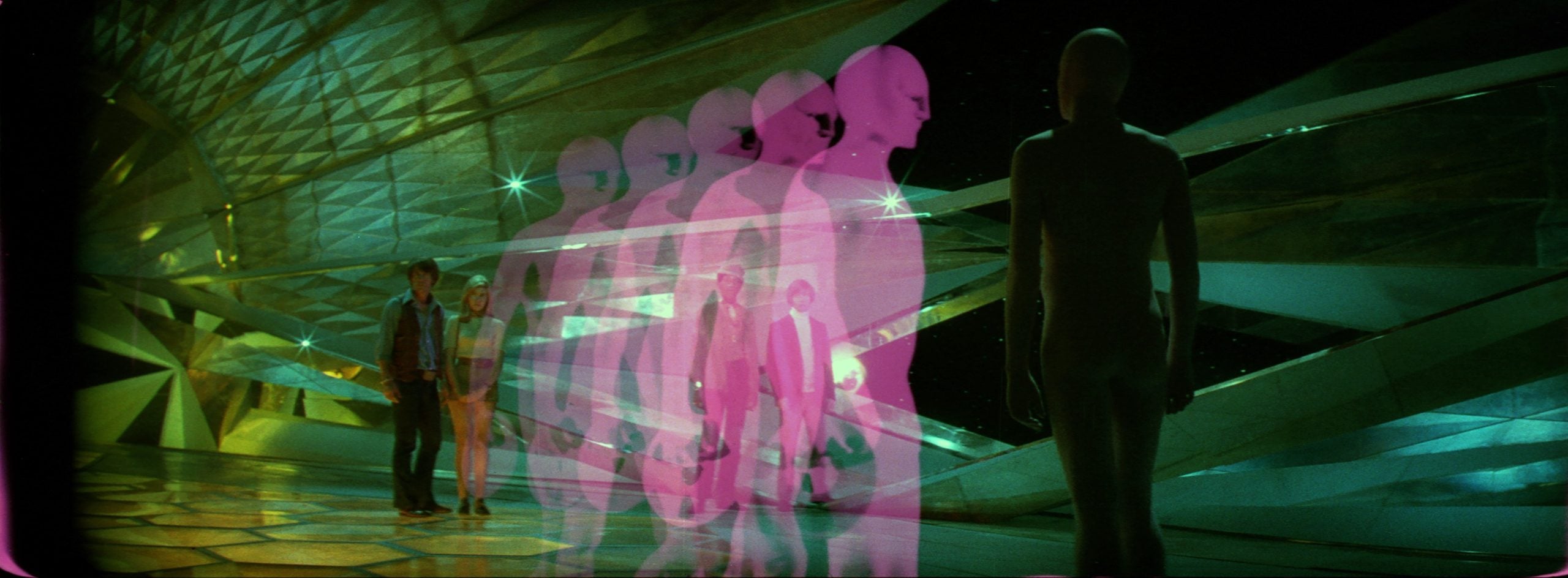 Two people stand in a futuristic, geometric room as translucent pink human figures walk toward a featureless mannequin-like figure.