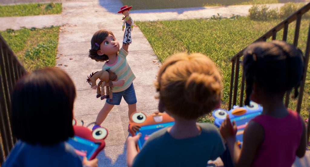 A young girl stands on a sidewalk holding up a cowboy doll while three other children holding toy steering wheels watch her.