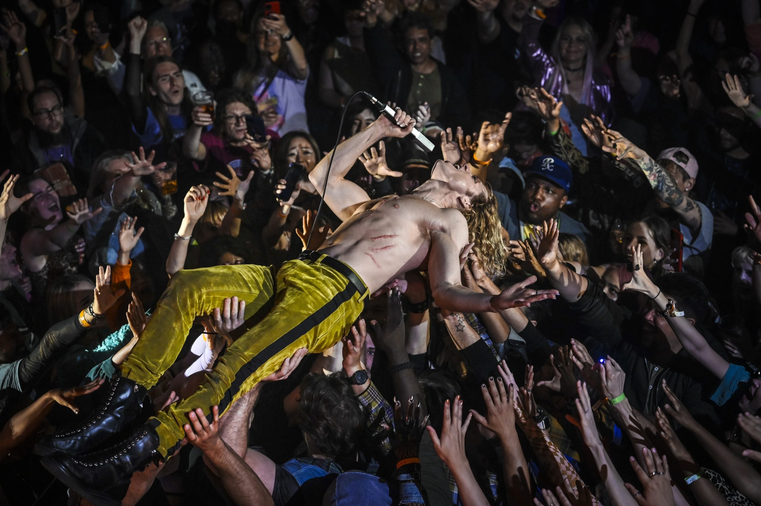 A shirtless performer in yellow pants crowd-surfs while singing into a microphone, surrounded by an enthusiastic audience reaching up toward him.