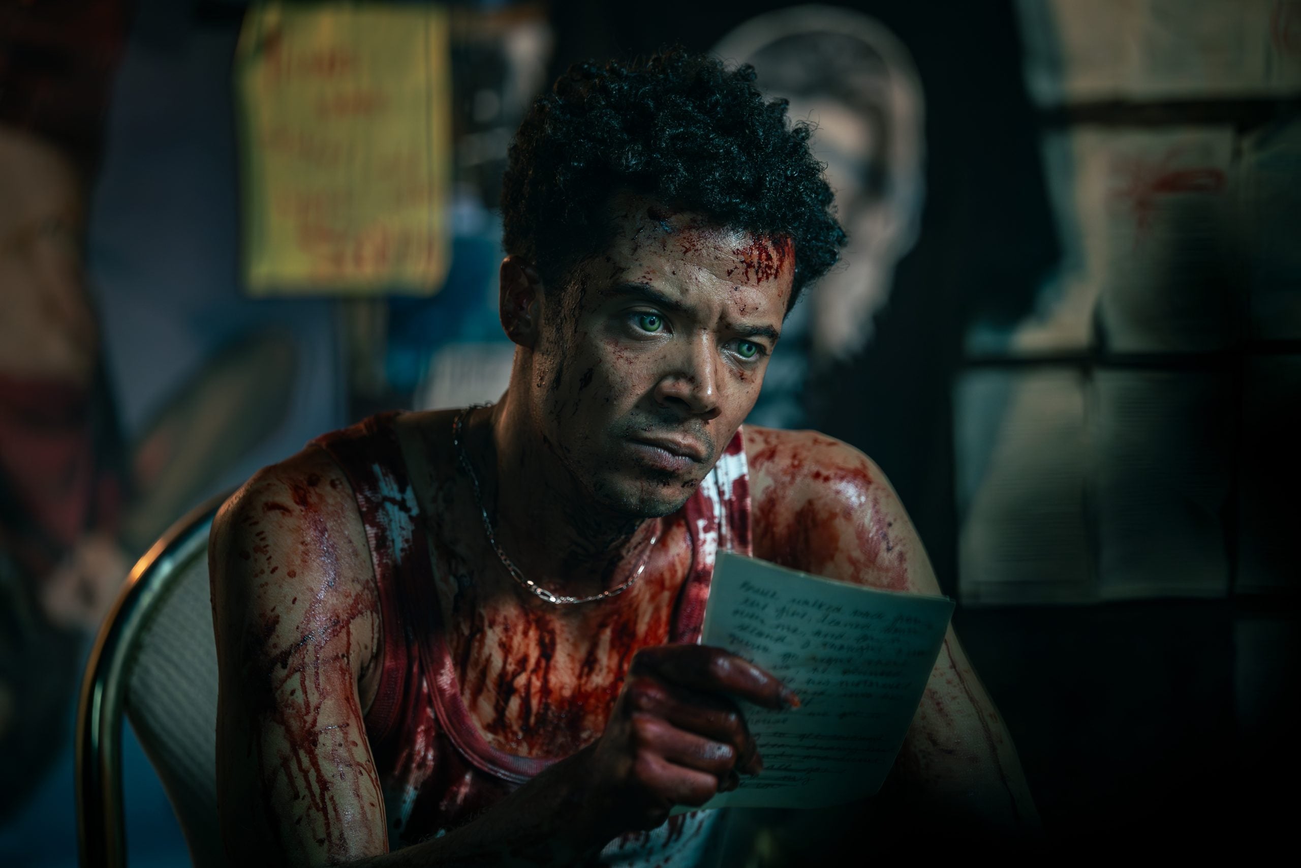 A man with bloodstained skin and clothing sits on a chair, holding a handwritten letter, looking intensely ahead in a dimly lit room with papers on the walls.